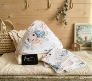 une cape de bain posée sur un meuble, la cape a comme motif des tortues bleues et des lingettes assorties