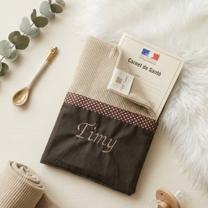 protège carnet de santé brun en bas et blanc cassé en haut.Il y a un biais chocolat au millieu et un prénom est brodé. Il est posé sur mon plan en bois avec un peu de déco