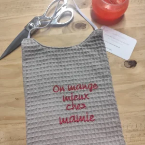 bavoir humour en nid d'ange d'un côté et de l'autre du coton.Il est posé sur mon établi et il y a un ciseaux et une boite orange.Il y a ; on mange mieux chez mamie brodé dessus