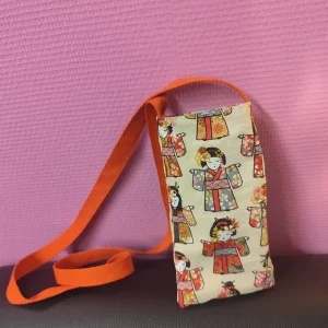 photo d'un sac téléphone en coton avec comme personnage une kokeshi qui est une poupée japonaise. Elle est posé sur un cuir noir et elle est cousue avec une sangle rouge