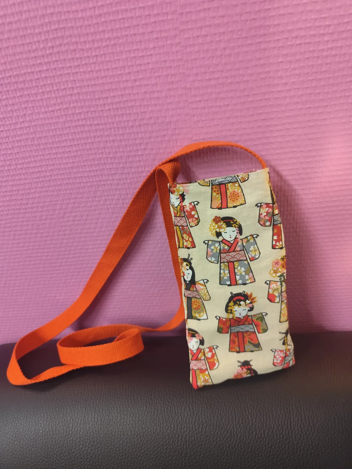 photo d'un sac téléphone en coton avec comme personnage une kokeshi qui est une poupée japonaise. Elle est posé sur un cuir noir et elle est cousue avec une sangle rouge