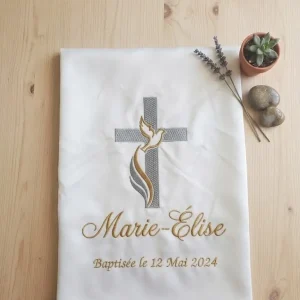 photo d'une écharpe blanche de baptême avec une croix grise et une colombe brodée. pliée sur mon établi ,il y a également le prénom et la date A côté de l'étole il y a une petite plante en déco