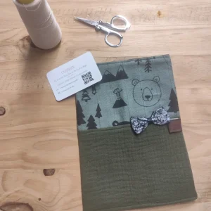 Photo protège carnet de santé vert uni sur le bas et dur le haut il y a des motifs .Un petit noeud est cousu devant au centre et il est posé sur mon établi
