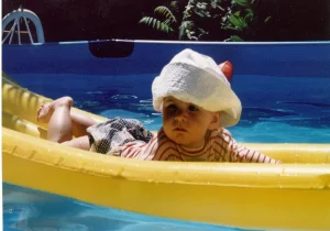 un bébé sur un matelas gonflable jaune installé dans une piscine. Elle a un joli chapeau blanc et un pull rayé