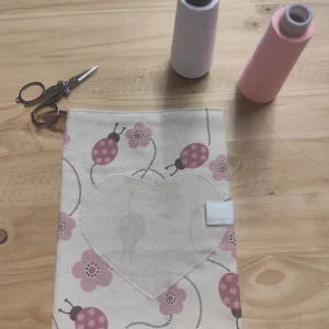photo d'un protège carnet de santé fabriqué dans du coton certifié gots.Ce sont de coccinelles roses avec des fleurs rose et un coeur cousu sur le davant .Posé sur mon établi en bois avec deux bibines de fils et un ciseaux