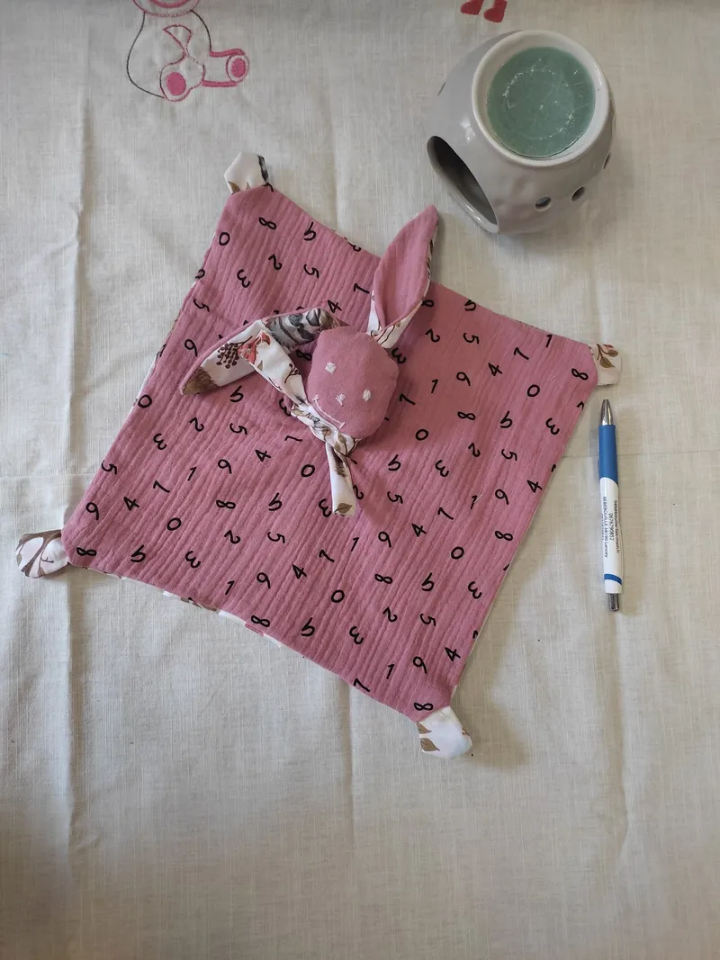 Un doudou en gaze de coton rose avec une tête de lapin avec des yeux brodés en blanc.Le gaze de coton est avec plein de petites lettres